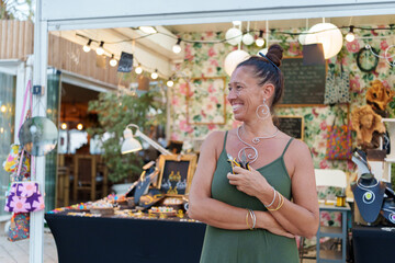 Mature woman proudly smiling, displaying handmade jewelry at colorful street market, highlighting entrepreneurial creativity and artisan skill