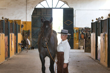 A young, attractive woman dressed in riding clothes and a traditional Spanish hat is standing in the stable next to her grey horse before setting off for a ride in the countryside.