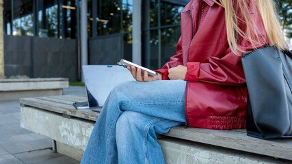 Obraz premium Woman checking her phone outdoors with a laptop nearby.
