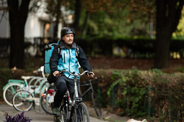 Fototapeta premium Delivery man riding bicycle wearing helmet and thermal backpack in city park