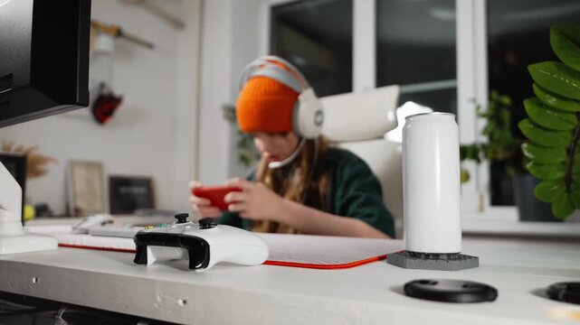 Teenage girl in orange beanie plays mobile game with red smartphone while wearing headset. Scene captures energy of Gen Z mobile gaming and cross-platform digital lifestyle at home.