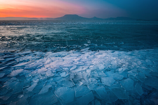 Fonyod, Hungary - Panoramic view of the frozen Lake Balaton on a cold winter afternoon with broken ice, golden colorful sunset sky and Badacsony mountain at background