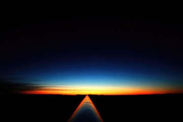 Dark landscape, water path leads to vibrant sunset horizon