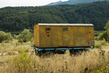 An old, yellow metal container, showing signs of rust and wear, is parked in a sun-drenched, dry grassy field. Behind it, a dense green forest covers rolling hills, creating a tranquil rural scene.