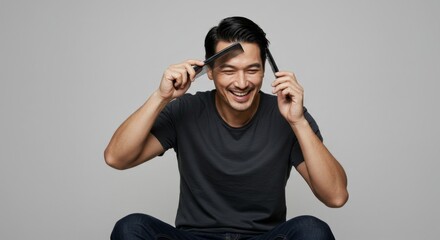 An asian man styling his hair with two combs, laughing with closed eyes. Happy guy caring for his appearance for barbershop promotion.