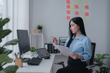 Young Asian businesswoman holding a pen and clipboard, analyzing statistical data on a tablet screen while working on a computer in a modern office environment
