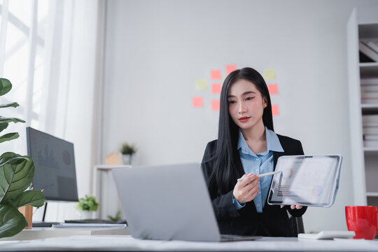 Young Asian businesswoman holding digital pen and using tablet presenting financial charts during online meeting via laptop in modern office - Powered by Adobe