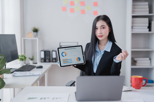 Asian businesswoman presenting sales performance chart on digital tablet during video conference with laptop in a modern office, analyzing data and discussing strategies for growth