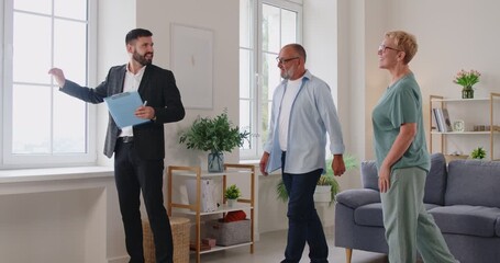 Realtor leads elderly clients on an apartment tour, discussing contract terms, purchase details in a bright living room. Real estate agent guides senior couple through apartment viewing.