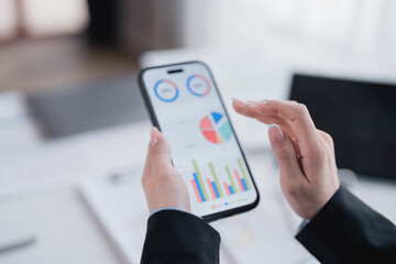 Businesswoman wearing a suit is analyzing financial charts and graphs displayed on a smartphone screen, indicating mobile business management and data analysis