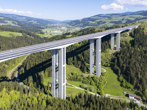 Aerial view of a modern, elevated highway cutting through a verdant valley, contrasting with the dark green forest and distant buildings, Wolfsberg, Carinthia, Austria.