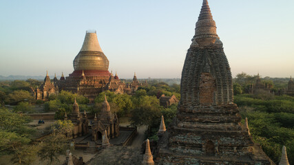 Aerial view of ancient temples rising majestically from the lush green canopy, their weathered brick contrasting with the soft morning light, Old Bagan, Mandalay Region, Myanmar.