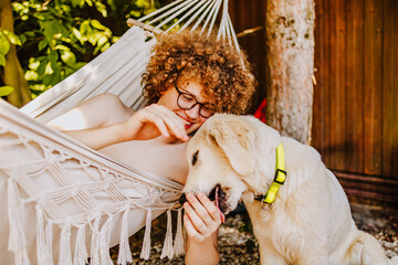 Teenager with curly hair relaxing in hammock playing with Golden Retriever