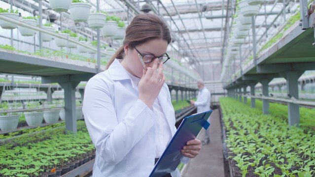 Female botanists in eyeglasses making notes on clipboard, working in greenhouse - Powered by Adobe