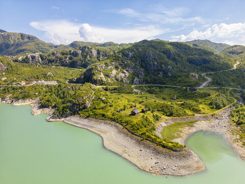 Aerial view of a winding road cuts through the verdant hills above the serene, turquoise Fjellhaugvatnet lake, reflecting the vast sky, Fjellhaugvatn, Matre, Vestland, Norway.