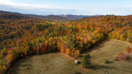 Aerial view of vibrant autumn foliage painting the hillsides in fiery hues, under a blue sky. A wooden cabin sits at the edge of a field and forest. White Mountains, New Hampshire, United States.