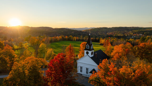 Aerial view of a white church with steeple, amidst a vibrant tapestry of red, yellow, and orange autumn trees with mountains beyond, under a golden sunset. White Mountains, NH, United States.