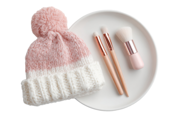 A pale pink and white knitted beanie sits atop a round white plate, alongside three makeup brushes