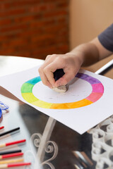 Child's hand erasing pencil marks on a color wheel drawing with a white eraser. Transparent surface with brushes. Natural light and copy space. Concept of visual learning and artistic development