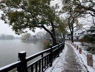 Naklejka premium Snowy scene of a lake path. A dark wooden fence lines a stone path leading towards traditional white buildings under a sky tinged with snow