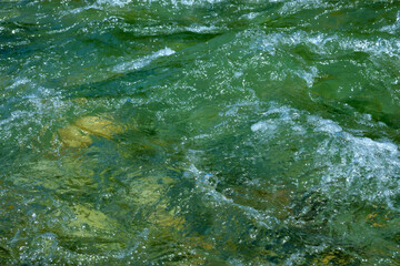 Close-up of greenish river water with swirling motion and white foam. The waves reflect sunlight, creating texture and sparkle. Capturing the energy, clarity and freshness of natural flowing water.