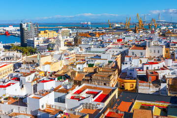 Obraz premium Aerial view shows a dense Cadiz cityscape of white buildings and a harbor. Cranes and a bridge are visible in the background, with the sea stretching to the horizon