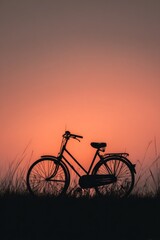 Obraz premium Silhouetted bicycle parked in grassy field at sunset. The sky fades from bright orange at the horizon to a soft pink above