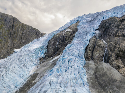 Aerial view of the icy blue glacier cascading down rugged, gray mountains under a cloudy sky, Buarbreen, Odda, Norway.