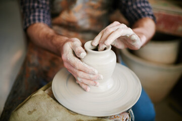 Caucasian young adult man shaping clay vase on pottery wheel, hands covered in wet clay, focusing on precise sculpting technique during ceramic creation process in workshop