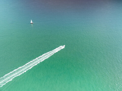 Aerial view of a motorboat cutting a white wake through the turquoise sea, while a lone sailboat drifts in the distance, Berni&Atilde;&uml;res-sur-Mer, Normandy, France.