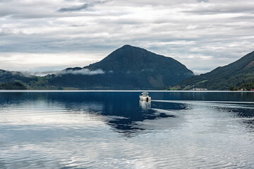 A boat moves quietly across still waters surrounded by majestic mountains and overcast skies, creating a peaceful atmosphere.