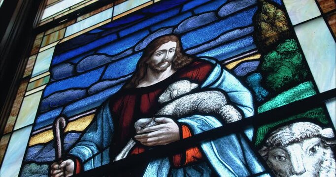 Vibrant stained-glass art inside a church sanctuary shows Jesus holding a lamb, surrounded by sheep, representing peace, faith, and religious tradition.