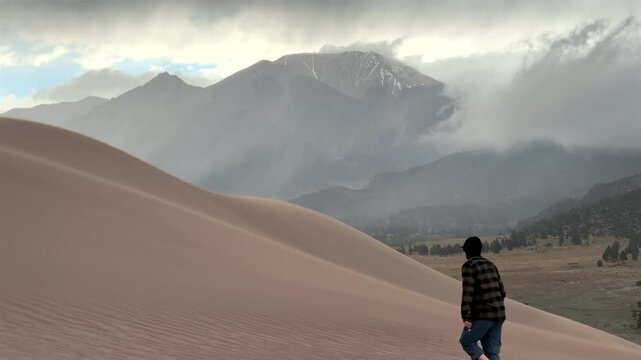 Stormy Great Sand Dunes National Park walking in windy sand sitting down Spring summer foggy cloudy raining mist Crestone Needle peak Colorado Sangre de Cristo range Rocky Mountains hills windy