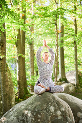 Woman practicing yoga outdoors in forest. Barefoot female on yoga mat surrounded by trees and large rocks, which suggests peaceful, natural environment ideal for meditation or yoga practice.