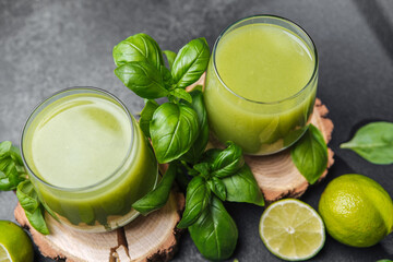 Two glasses of green juice with basil leaves and limes on wooden slices against dark backdrop