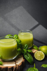 Two glasses of green smoothie with fresh basil and lime on dark background
