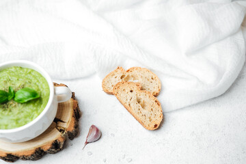 Healthy green soup with basil and bread slices on a white textured surface with cloth