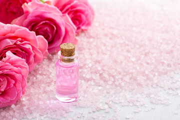 Bottle with rose oil, spa salt crystals and pink roses  on white wooden table.