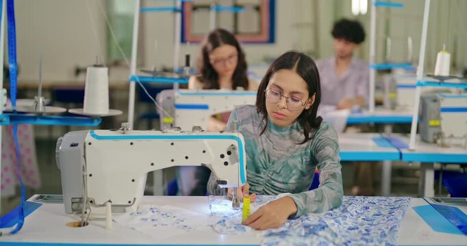 Indian asian young adult gen z student lady wear glass and measure tape around neck sitting class room learn self sewing machine focused serious girl do fashion design course indoor college place