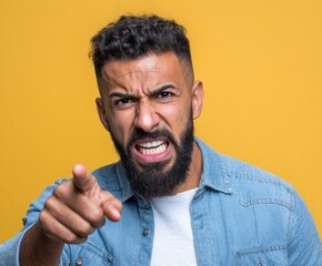 Fototapeta premium A man with an intense expression, pointing directly at the viewer, projects anger and accusation against a bright yellow background.