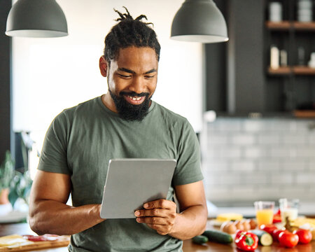 Happy young afro american man having fun preparing food and looking for recipes online using a tablet in kitchen, or a young businessman working from home office making connections online