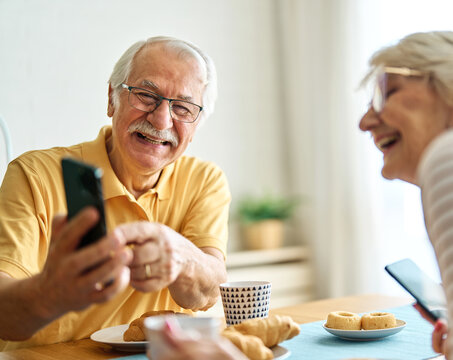Portrait of an elderly senior couple having breakfast and using a smart phone at home. Happy healthy affectionate senior couple talking and showing smartphone screen to one another and texting and sit