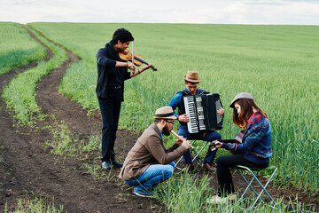 Group of young adult multiethnic musicians performing outdoors in green field, standing and sitting while playing violin, accordion, guitar and flute, focused on instruments