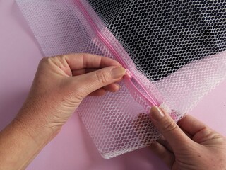 Closeup of woman hands zipping a white mesh laundry bag up. Woman hands closing a laundry bag zipper on pink background.