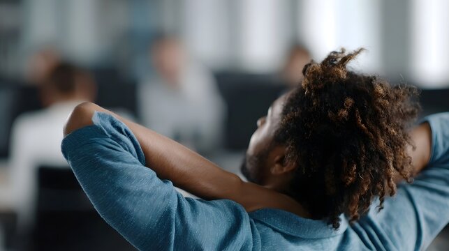 Office worker enjoys a moment of relaxation leaning back in a chair with hands clasped behind head blurred background with colleagues