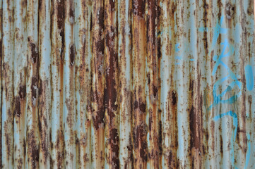 Rusty corrugated metal sheet with peeling paint and traces of blue graffiti, creating an urban industrial texture background.
