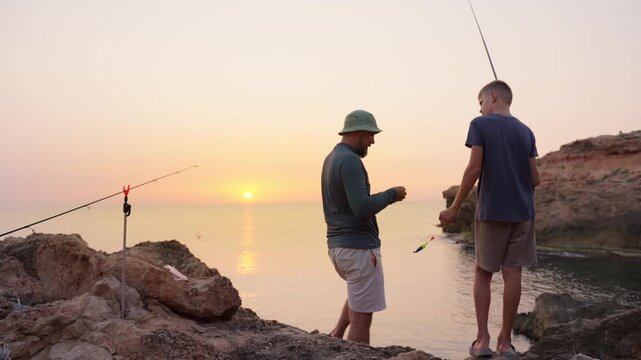 Father and son preparing fishing rods together on rocky coastal shoreline during golden sunrise, sharing quiet moment of family connection and outdoor leisure