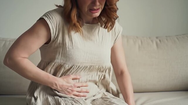 Woman Suffering from Abdominal Pain Sitting on Light Beige Sofa in Softly Lit Room Gripping Stomach in Agony Light Dress Health Issues and Discomfort Sickness Nausea Discomfort