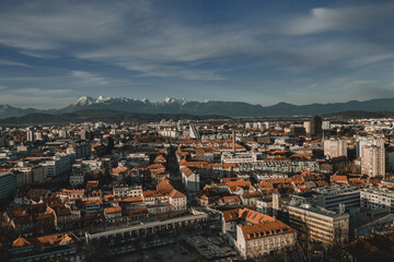 Obraz premium A panoramic view of the city of Ljubljana, with its red roofs and historic architecture, set against the majestic, snow-capped Julian Alps. 