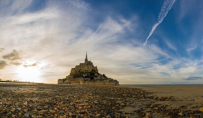 Panoramic view of Mont-Saint-Michel at sunset. Historic abbey on a rocky island surrounded by tidal sands and a dramatic sky. Iconic French landmark and UNESCO heritage site.